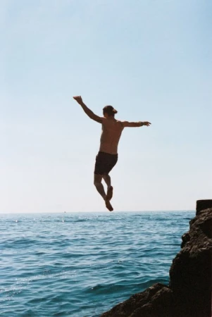 Man jumping off a rocky cliff into the blue ocean captured mid-air by photographers AnaÏs and Dax.