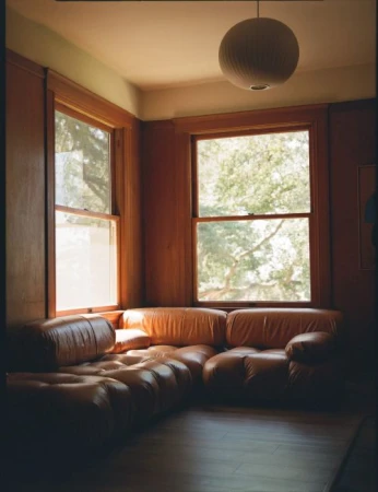 A brown leather sectional sofa positioned in the corner of a wood-paneled living room with large windows and a round pendant light.