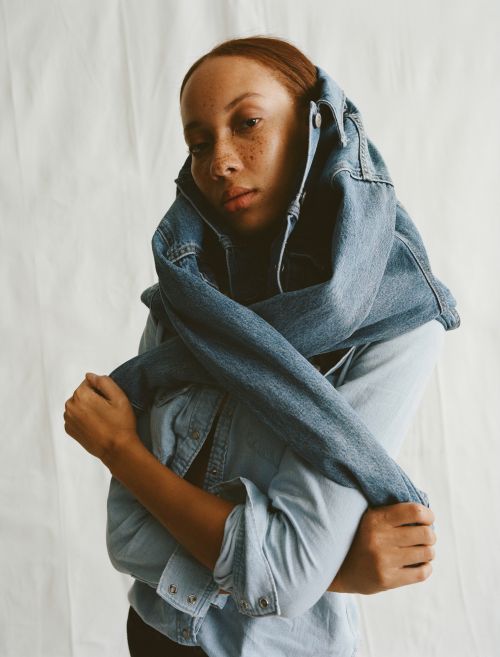 Portrait of woman with auburn hair and freckles, posing indoors wrapped in a blue jean jacket against a light neutral background.