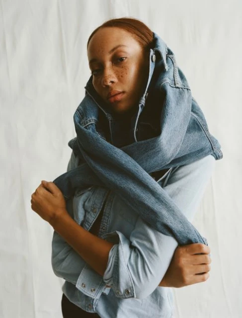 Portrait of woman with auburn hair and freckles, posing indoors wrapped in a blue jean jacket against a light neutral background.