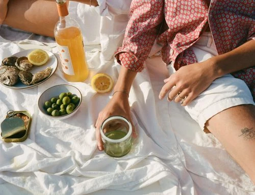 Outdoor picnic blanket still life featuring oysters, olives, lemons, a green drink, and a bottle of orange liquid in the sunlight.