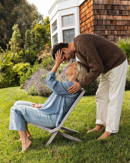 A couple embrace outdoors in a sunlit garden, the woman seated on a wooden chair and the man standing, with trees and a house behind them.