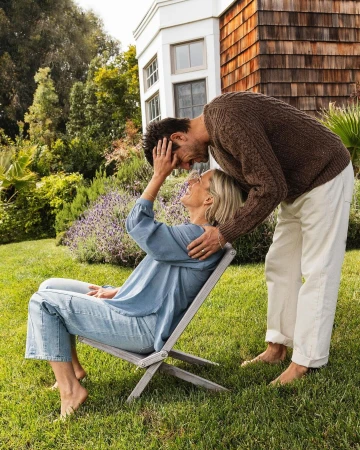 A couple embrace outdoors in a sunlit garden, the woman seated on a wooden chair and the man standing, with trees and a house behind them.