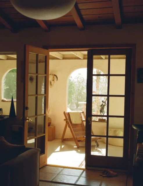 View from inside a home to an outdoor patio with an open glass door and a wooden lounge chair in the sunlight.