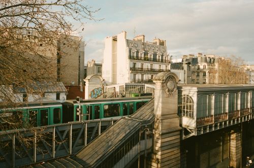 Urban scene of a green train on an elevated track in Paris, France, with buildings in the background and bare trees in the foreground.
