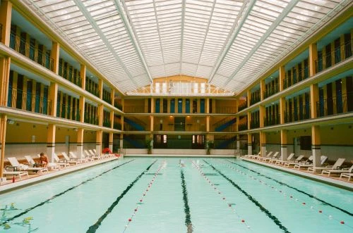 Indoor swimming pool with person resting on lounge chair; multi-level balconies and a high, translucent roof.