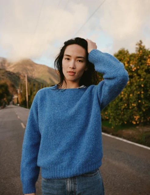 A woman in a blue sweater stands on a road in a scenic outdoor setting with mountains in the background, looking directly at the camera.