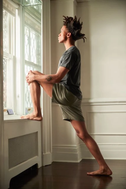 Adult male with dreadlocks stretches his leg on top of a white radiator beneath a window inside a sunlit home with dark wood floors.