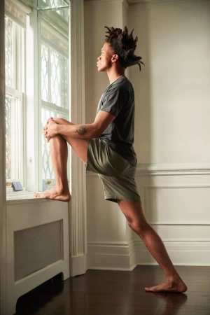 Adult male with dreadlocks stretches his leg on top of a white radiator beneath a window inside a sunlit home with dark wood floors.
