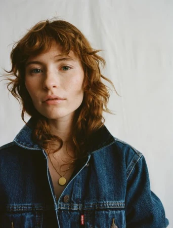 Portrait of a young adult female with red wavy hair wearing a denim jacket and gold necklace against a white background.