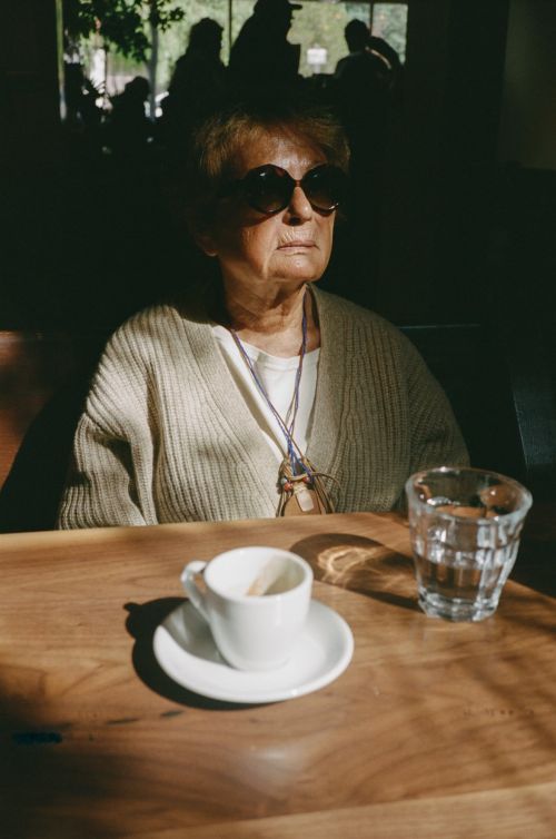 Elderly woman in sunglasses sits at a wood table in a cafe with a coffee cup and glass of water, bathed in sunlight, shadowed background.