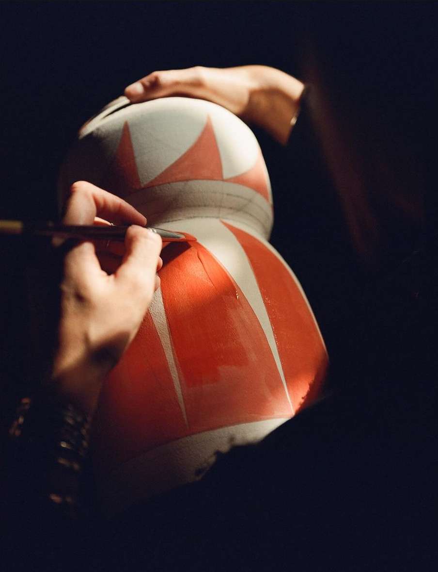 Artist painting red geometric shapes on a large white vase. Focus on the artist's hands holding the brush, and the vase itself.