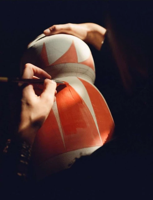 Artist painting red geometric shapes on a large white vase. Focus on the artist's hands holding the brush, and the vase itself.