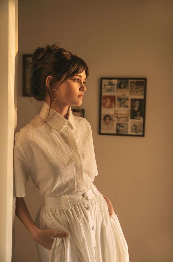 Young woman in white dress leans against wall indoors, featuring natural light, hoop earrings, and a photo gallery backdrop.