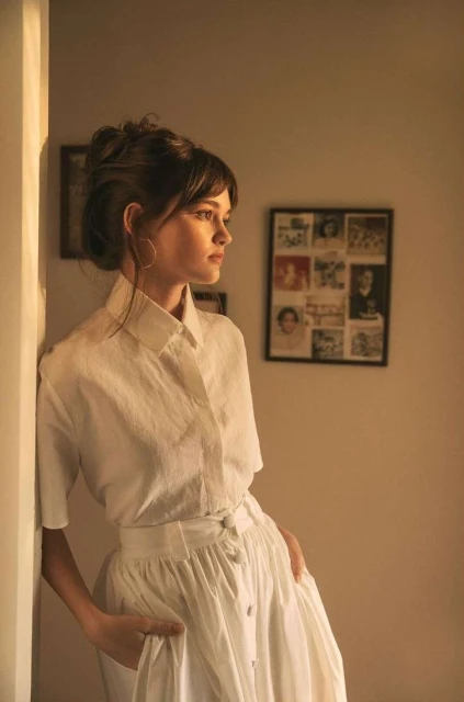 Young woman in white dress leans against wall indoors, featuring natural light, hoop earrings, and a photo gallery backdrop.