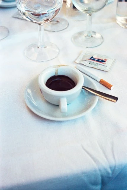 Overhead shot of a cup of espresso on a saucer with a cigarette and sugar packet on a white tablecloth, with clear glasses in the background.