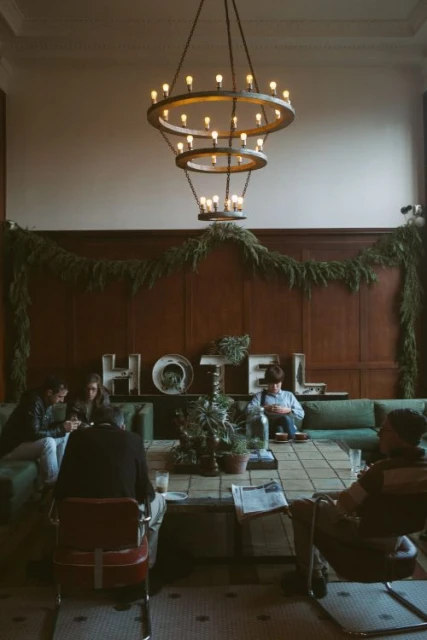 People gather in a hotel lobby with a large chandelier and plants. Guests sit on sofas and chairs, creating a relaxed atmosphere.