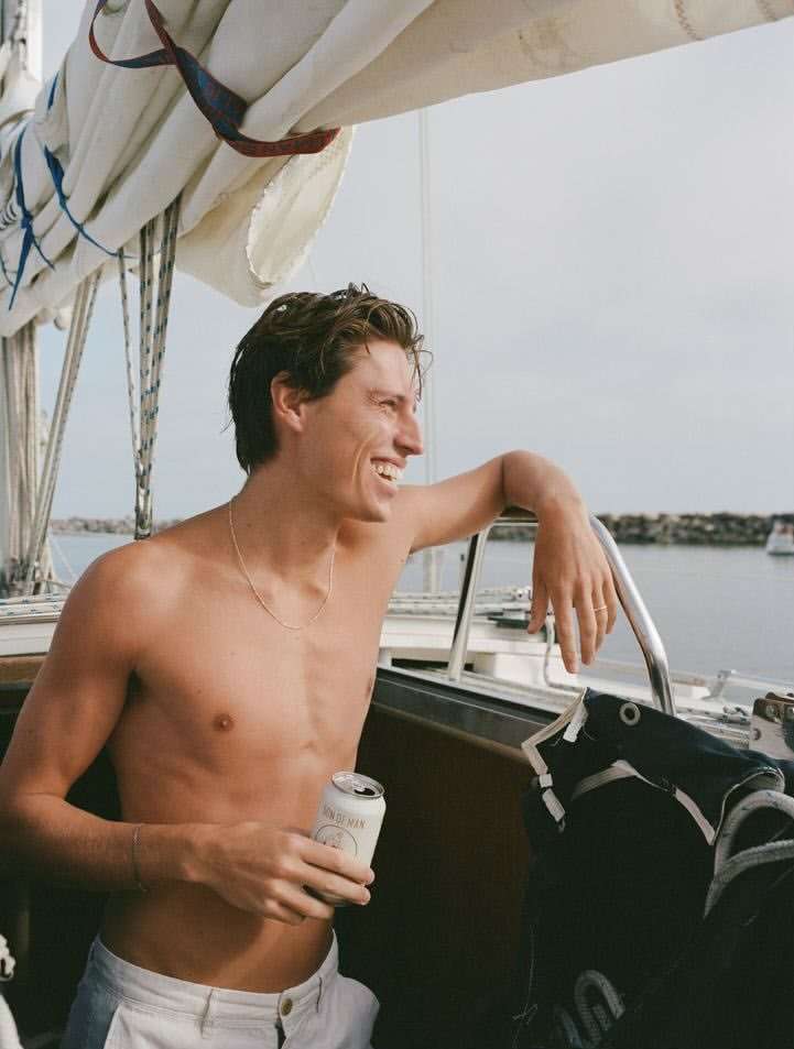 Smiling shirtless man holds a drink on a sailboat, enjoying a sunny day on the water with sails, water, and the horizon in the background.