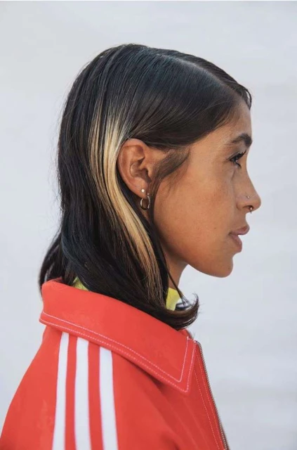 Profile portrait of an adult female with black hair and a blonde streak, wearing a red Adidas track jacket against a white background.