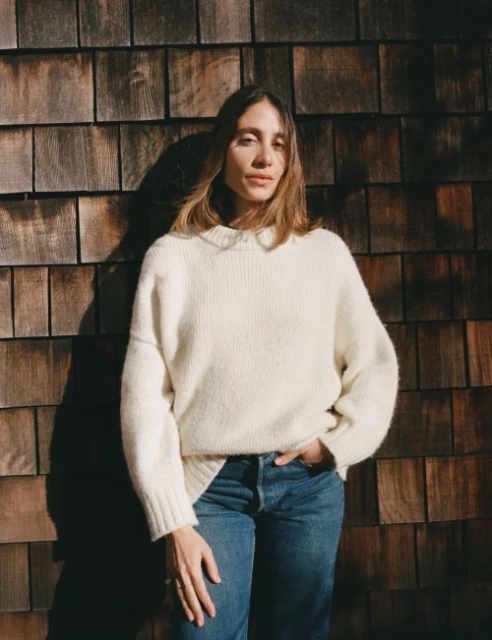 A woman in a cream-colored sweater and blue jeans poses against a rustic wood-paneled wall with soft lighting and natural earthy tones.