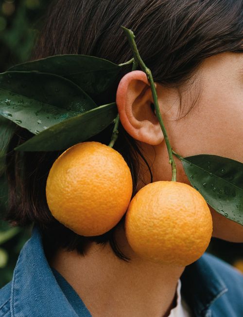 Close-up shot of a person with two oranges hanging near their ear, alongside green leaves and wearing a denim jacket.