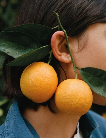 Close-up shot of a person with two oranges hanging near their ear, alongside green leaves and wearing a denim jacket.