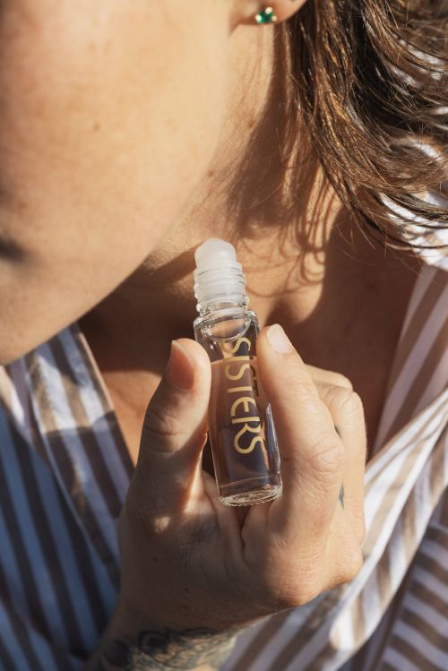 Close-up of a woman applying a glass rollerball bottle of Sisters beauty product to her neck, wearing a striped shirt.
