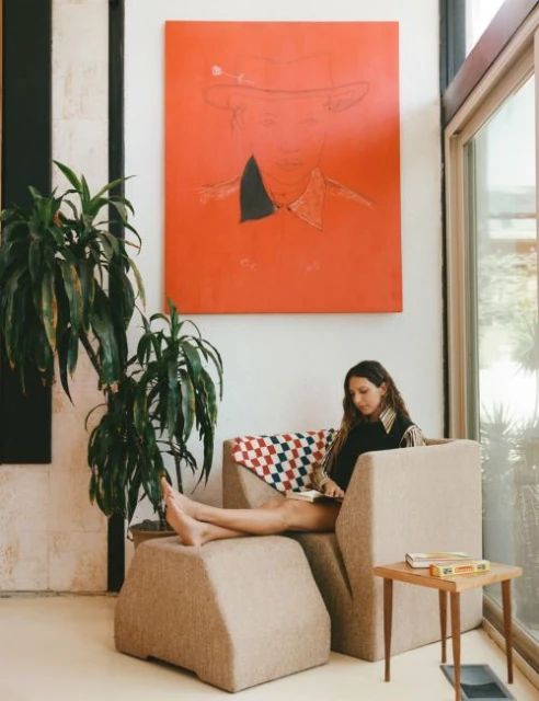 Woman relaxing barefoot in a beige chair in a brightly lit room with a vibrant orange painting, potted plant, and small side table.