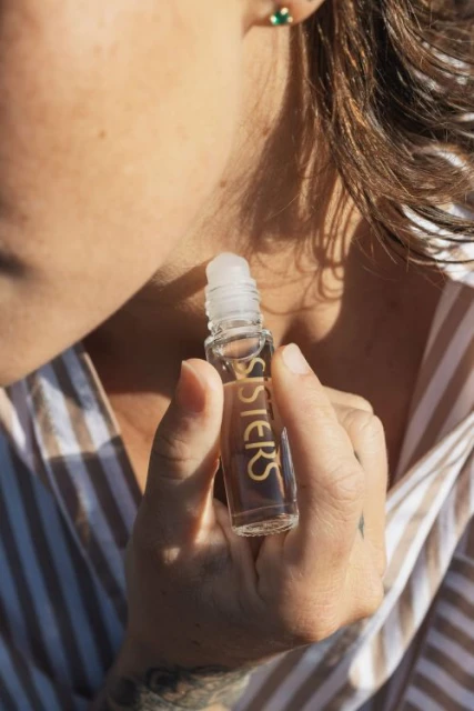 Woman applying Sisters aromatherapy oil to her neck. Close-up shot of the hand holding a clear glass bottle with gold lettering.