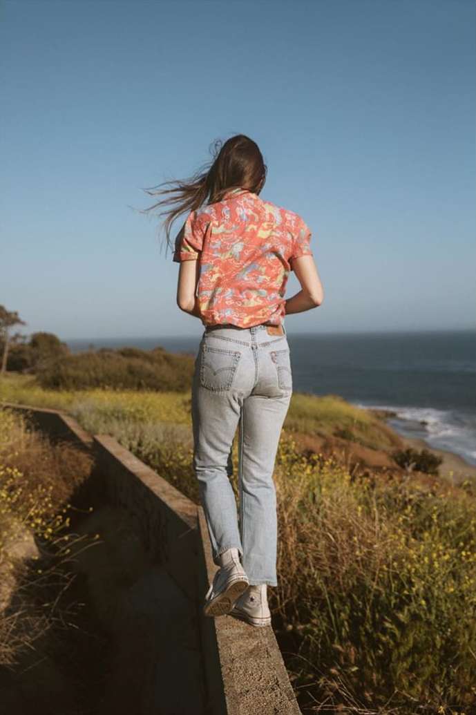 A woman walks along a concrete wall near the ocean, wearing a patterned top and jeans with her back to the camera on a sunny day.