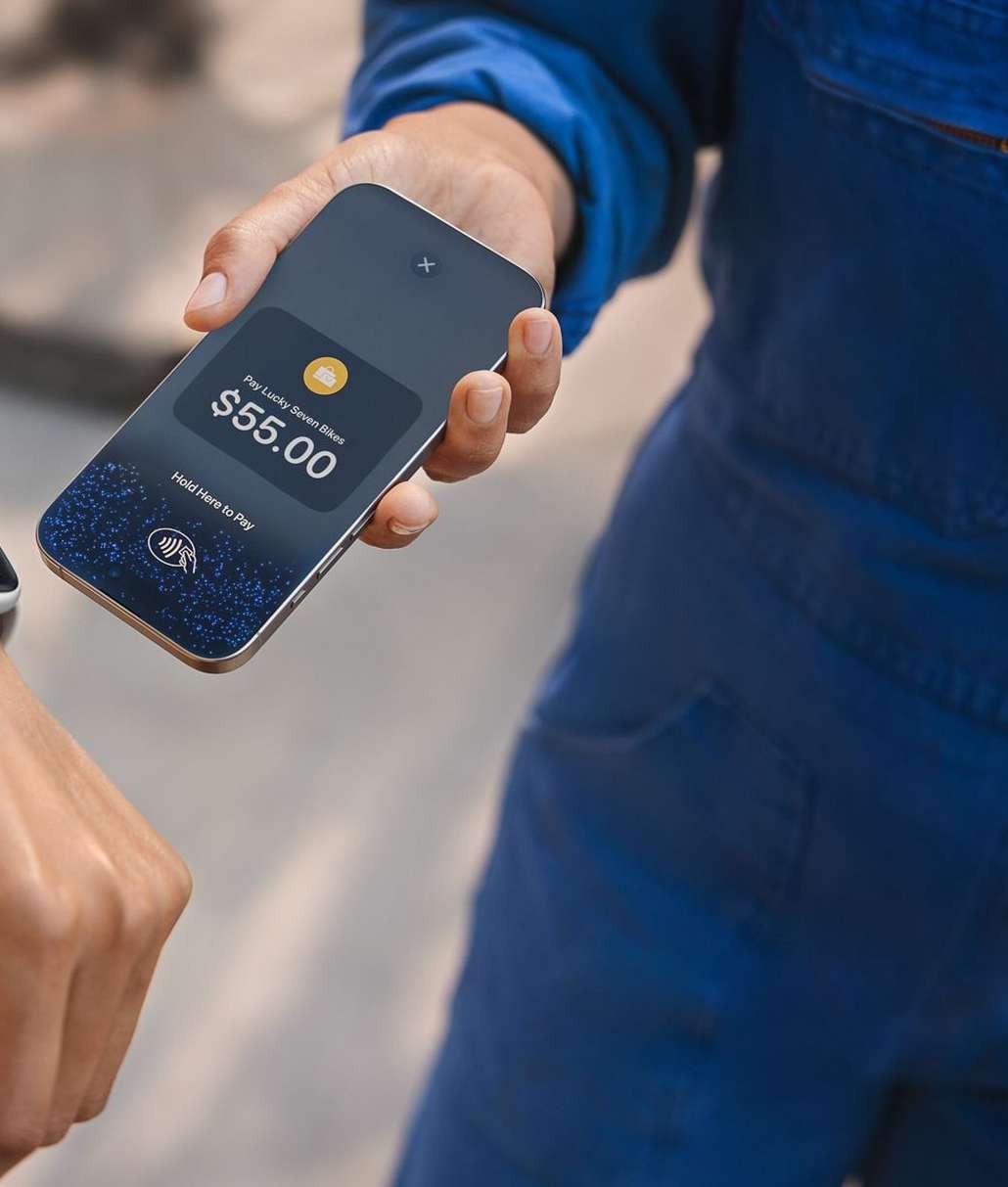 Person holds a smartphone showing a payment screen with $55.00 while another person extends a smartwatch for contactless payment.