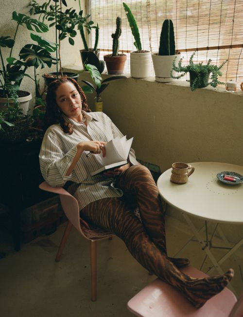 Adult woman with patterned pants sitting in a pink chair reading a book surrounded by potted plants in an indoor setting, daytime.