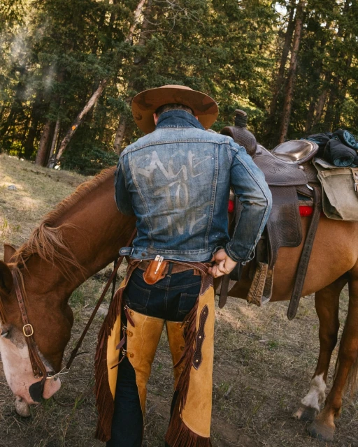 A horse stands in a forest as a person wearing a cowboy hat, jean jacket, and chaps adjusts their belt near the animal.