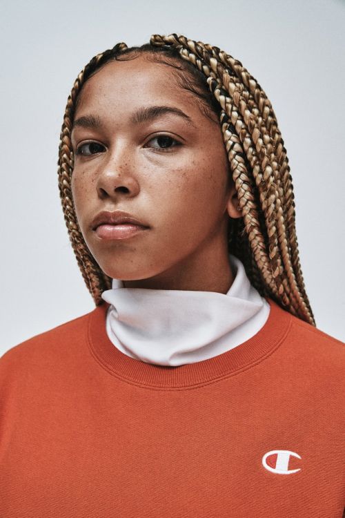 Portrait of a young adult female with braided hair wearing a Champion sweatshirt, set against a neutral background in a studio.