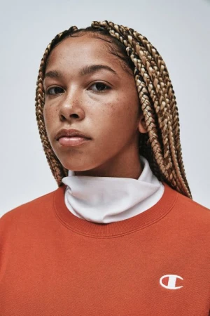 Portrait of a young adult female with braided hair wearing a Champion sweatshirt, set against a neutral background in a studio.