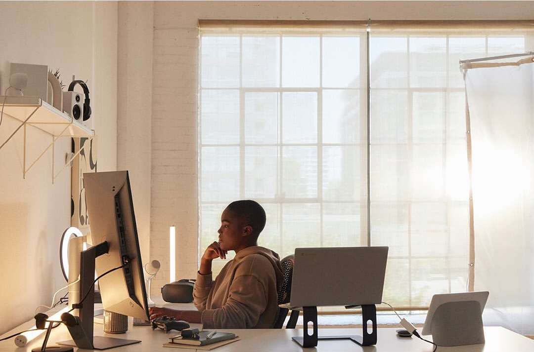 A woman with a short haircut sits at a white desk with a computer, laptop, and VR headset in a brightly lit indoor room.