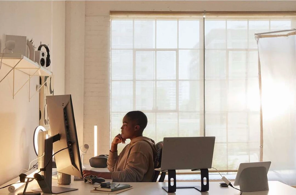 A woman with a short haircut sits at a white desk with a computer, laptop, and VR headset in a brightly lit indoor room.