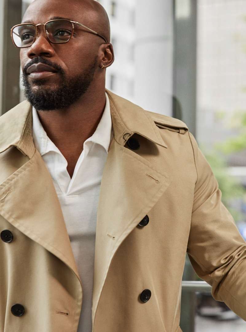 Close-up shot of a bald African American male wearing glasses and a trench coat, looking thoughtfully to the side, in natural lighting.