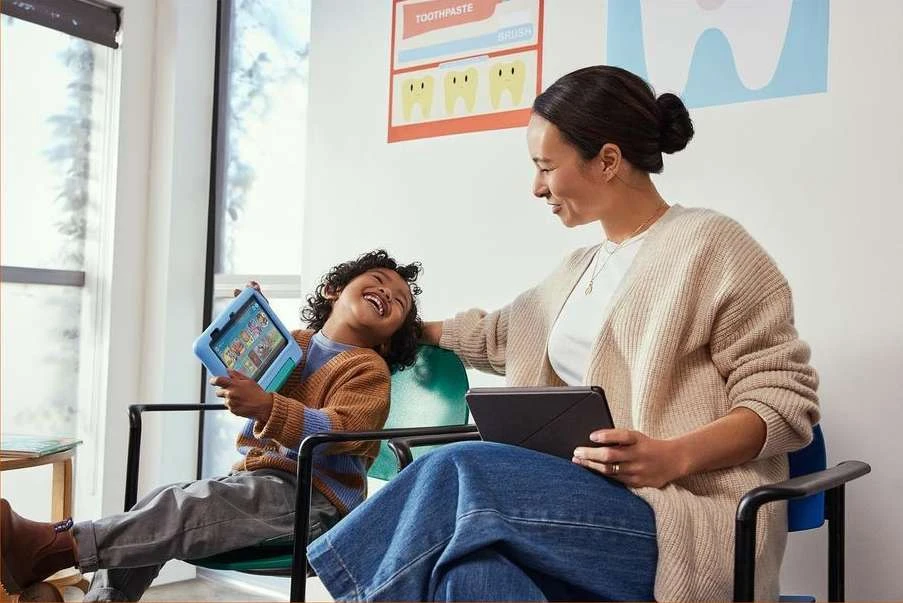 Smiling child on chair with tablet as mother looks on, both at doctor's office, capturing a sweet family moment.
