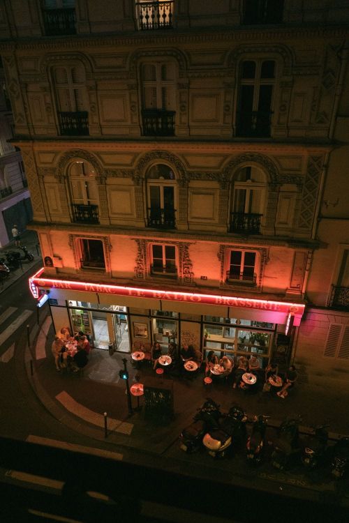 Outdoor cafe scene at night, featuring people seated at tables, illuminated by warm, orange lighting from building fixtures.