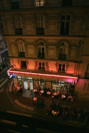 Outdoor cafe scene at night, featuring people seated at tables, illuminated by warm, orange lighting from building fixtures.