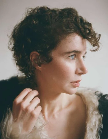 Portrait of a woman with short curly brown hair and blue eyes wearing a textured fluffy collar against a light background.