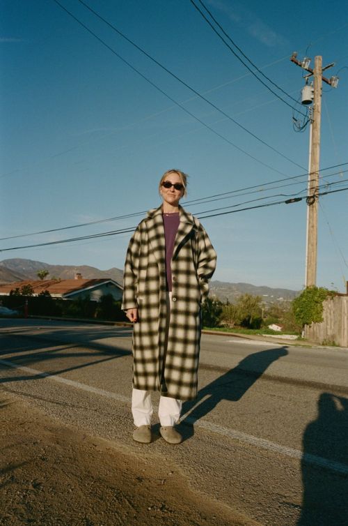 Adult female in a patterned coat and sunglasses standing on an asphalt road with a residential neighborhood and mountains in the background.