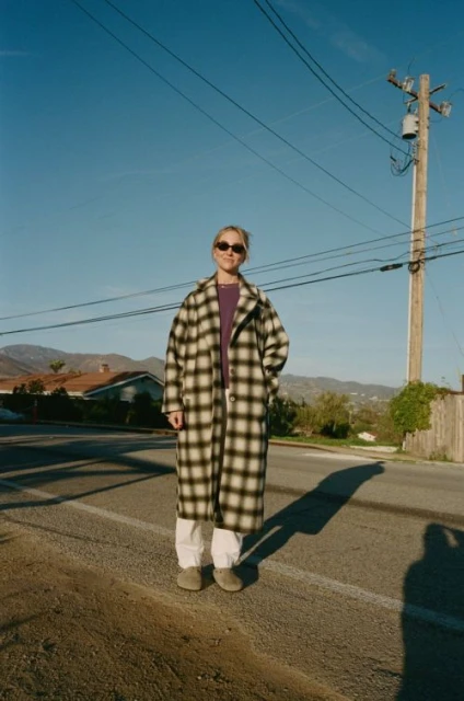 Adult female in a patterned coat and sunglasses standing on an asphalt road with a residential neighborhood and mountains in the background.