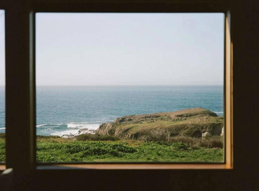 Scenic view of the ocean and coastline through a window frame, showing a grassy headland and clear sky, capturing a serene landscape.