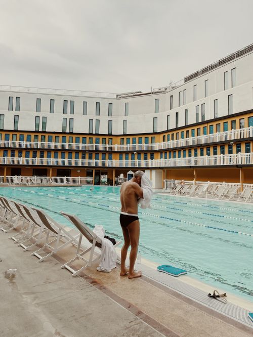 Man drying off with a towel after swimming at an outdoor pool with rows of empty lounge chairs and an apartment building backdrop.
