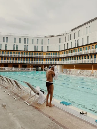 Man drying off with a towel after swimming at an outdoor pool with rows of empty lounge chairs and an apartment building backdrop.