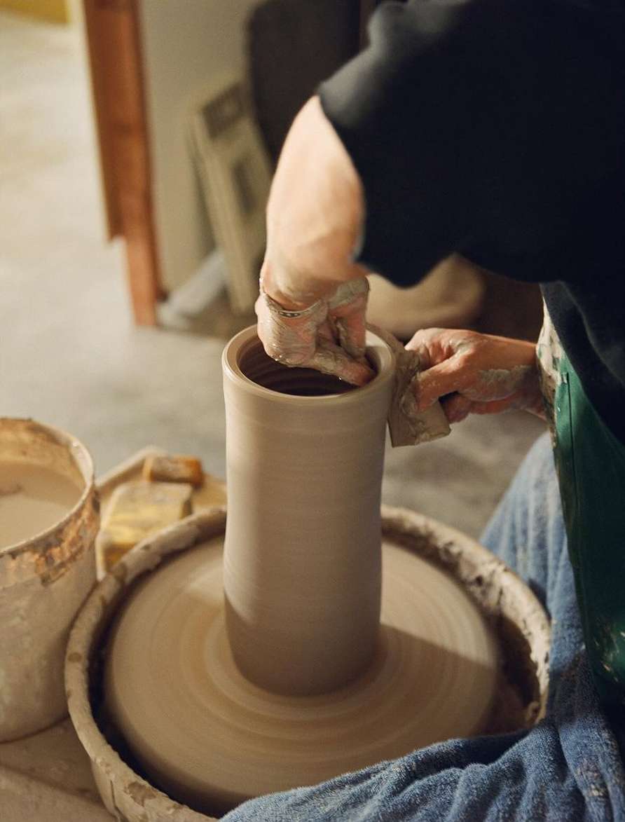 Person shapes clay on a spinning pottery wheel in an indoor workshop, crafting a tall cylinder-shaped pot. Soft lighting highlights the process.