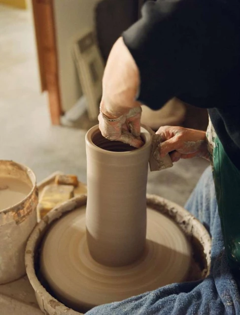 Person shapes clay on a spinning pottery wheel in an indoor workshop, crafting a tall cylinder-shaped pot. Soft lighting highlights the process.
