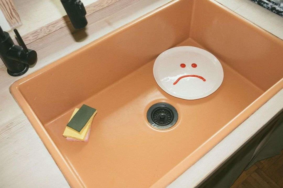 Close-up of an orange kitchen sink featuring a plate with a sad face drawn on it, alongside dish sponges and a modern black faucet.